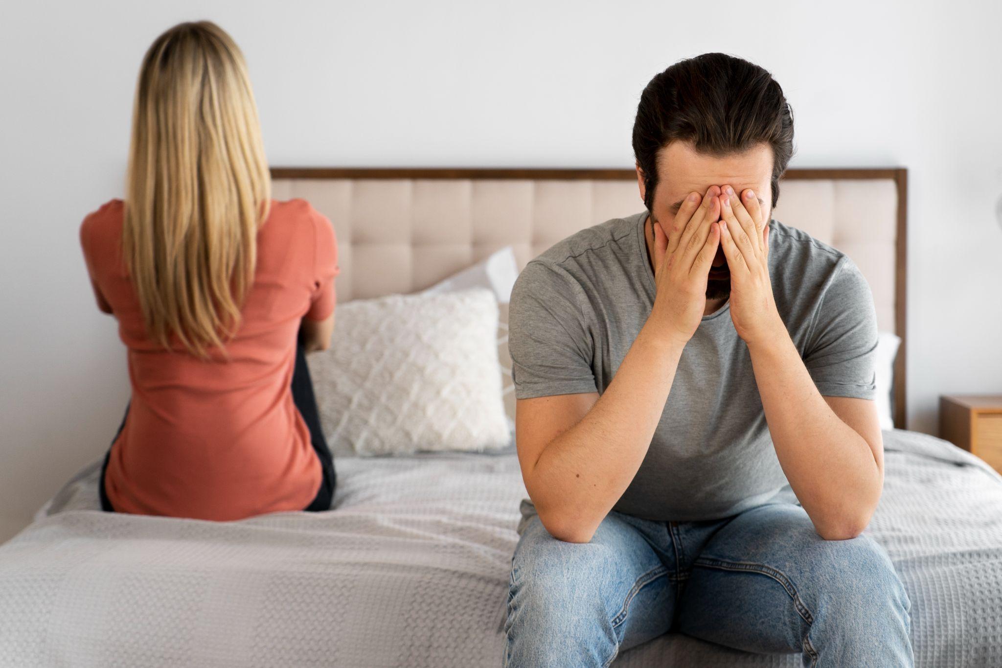 A man and woman sitting apart on a bed, looking distressed and upset.