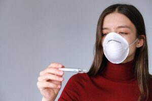 Woman wearing a mask checking temperature with thermometer while learning how to reduce high fever safely.