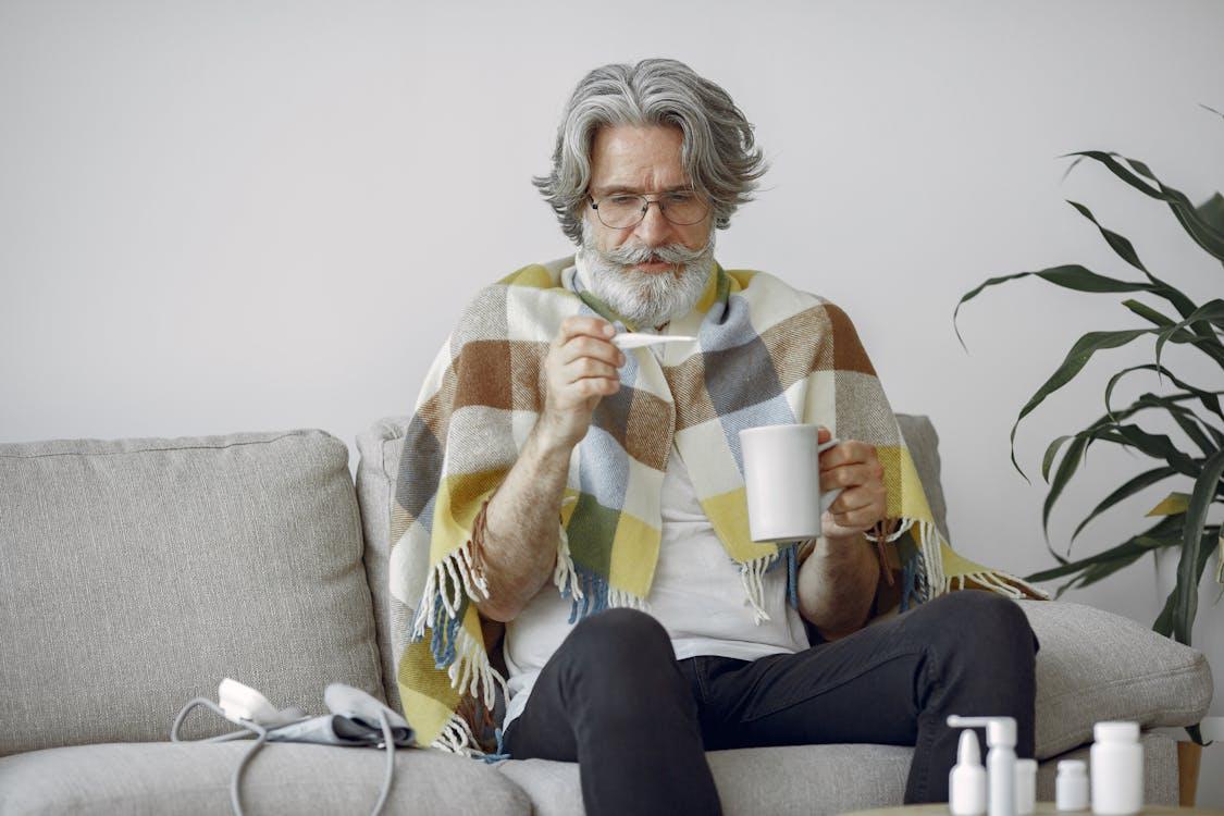 An elderly man checking his temperature while wrapped in a blanket on a couch